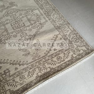 Peut inclure: Tapis beige et marron &agrave; motifs g&eacute;om&eacute;triques. Le tapis pr&eacute;sente un motif central avec des formes r&eacute;p&eacute;t&eacute;es et une bordure avec un design d&eacute;taill&eacute;. Le tapis a une bordure &agrave; franges et le texte "NAZAF CARPET" est visible.