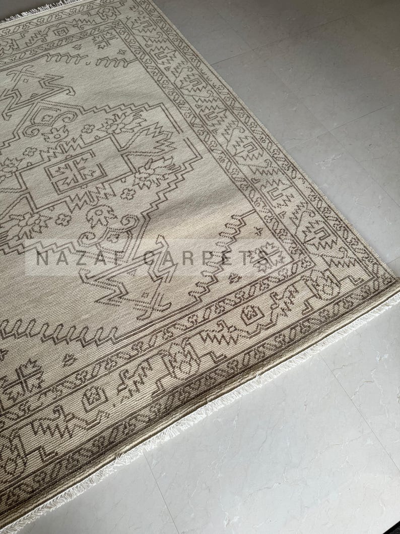 Peut inclure: Tapis beige et marron &agrave; motifs g&eacute;om&eacute;triques. Le tapis pr&eacute;sente un motif central avec des formes r&eacute;p&eacute;t&eacute;es et une bordure avec un design d&eacute;taill&eacute;. Le tapis a une bordure &agrave; franges et le texte "NAZAF CARPET" est visible.