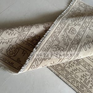 Peut inclure: Tapis beige et marron &agrave; motifs, avec une section enroul&eacute;e r&eacute;v&eacute;lant le dessous. Le tapis pr&eacute;sente un motif g&eacute;om&eacute;trique et une bordure &agrave; franges. La palette de couleurs est neutre, avec des nuances de beige et de marron.