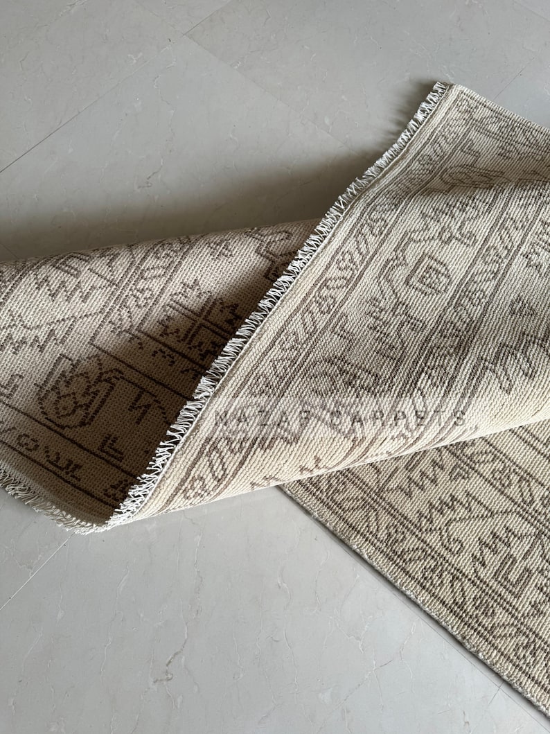 Peut inclure: Tapis beige et marron &agrave; motifs, avec une section enroul&eacute;e r&eacute;v&eacute;lant le dessous. Le tapis pr&eacute;sente un motif g&eacute;om&eacute;trique et une bordure &agrave; franges. La palette de couleurs est neutre, avec des nuances de beige et de marron.