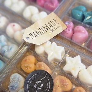 May include: A clear plastic container filled with various scented wax melts. The wax melts are in the shapes of hearts and stars. The container has a label that says "Handmade with love..." and another label that says "Oh My Scents" with a cruelty-free logo.