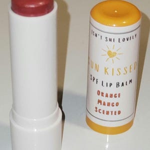 Könnte beinhalten: Zwei Lippenbalsam-Tuben. Eine ist weiß mit rotem Deckel, die andere ist weiß mit orangem Deckel. Die Tube mit dem orangen Deckel trägt den Text "Isn't She Lovely" und "Sun Kissed SPF Lip Balm Orange Mango Scented".