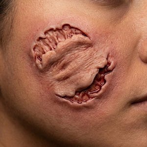 Pode incluir: Uma prótese de maquilhagem aplicada numa bochecha, com uma textura de pele circular e elevada, lacerações profundas e irregulares em tons de vermelho e detalhes de tecido exposto, concebida para simular uma ferida realista.