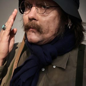Può includere: Un casco verde oliva scuro con occhiali protettivi cromati, occhiali tondi con montatura in metallo, un folto baffo marrone e una sciarpa blu navy. L'abbigliamento include una camicia oliva abbottonata con bretelle e un anello d'argento.