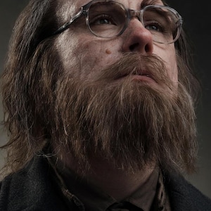 May include: A portrait of a person with long, wavy brown hair and a full, thick brown beard. The person wears clear-framed glasses and a dark charcoal coat over a brown collared shirt. The lighting is soft, highlighting facial features.