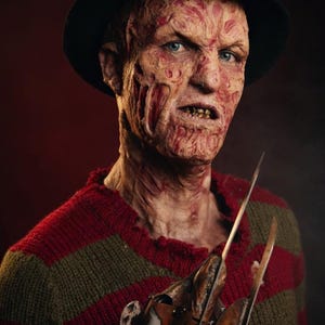 Op de afbeelding: Een personage met een zwarte fedora en een rood en olijfgroen gestreepte trui. Het personage heeft een prothetisch gezicht met een getextureerde, littekenachtige huid, zichtbare tanden en blauwe ogen. Een metalen handschoen met lange messen.