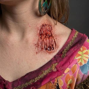 May include: A realistic prosthetic wound appliance applied to the collarbone area, featuring deep red lacerations and textured skin effects. The garment is a distressed pink floral fabric with gold trim, paired with a large rainbow peace sign earring.