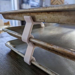 May include: A white plastic baking sheet rack supporting two metal baking sheets. The rack is designed to hold the sheets in place, with the sheets resting on the metal bars. The rack is on a dark wooden surface.