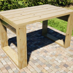 May include: A simple wooden table with a rectangular top and a square base. The table is made of light-coloured wood and has a natural finish. It is sitting on a patio made of small, irregularly shaped stones.
