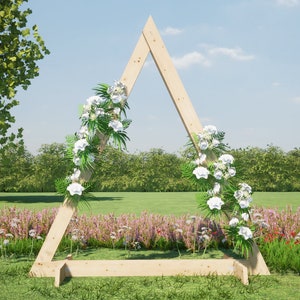 Puede incluir: Un arco triangular de madera decorado con flores blancas y vegetación. El arco está situado en un campo de hierba con una fila de árboles en el fondo.