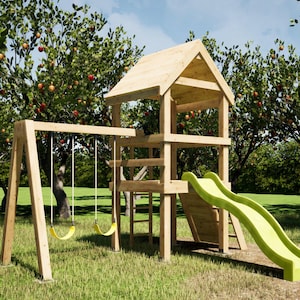 May include: A wooden playset with a swing set, slide, and playhouse structure. The playset is made of light-colored wood and has a green slide. The swings are yellow. The playset is in a grassy area with trees in the background.
