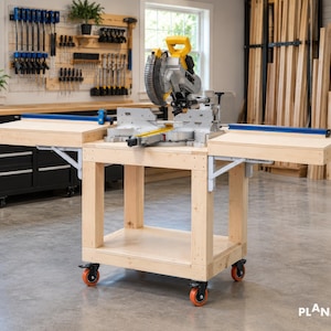 Peut inclure: Une station de scie à onglets en bois clair comprenant une plateforme centrale avec deux ailes latérales pliables, une étagère de rangement inférieure et quatre roulettes verrouillables. Une scie jaune et grise est fixée sur le dessus.