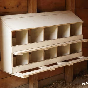 Puede incluir: Un gallinero de madera con dos niveles de ponederos. El gallinero tiene un tejado inclinado y está montado sobre una estructura de madera. El color natural de la madera se integra en el entorno del granero.