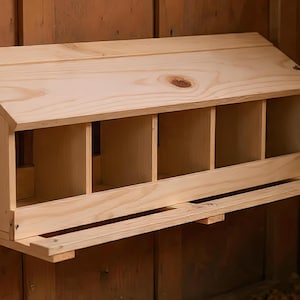 Puede incluir: Un nido de madera con techo inclinado y múltiples compartimentos. La caja es de madera de color claro y está montada en una pared de madera. Tiene una pequeña percha que se extiende desde el frente.