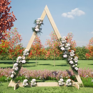 May include: A wooden triangular archway decorated with white flowers and greenery, standing in a grassy field with trees in the background.