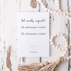 Könnte beinhalten: Eine weiße Karte mit goldener Schrift, auf der "Ich mach's einfach! Ich informiere vorher! Ich informiere vorher!" steht, mit einem goldenen Dreiecksymbol und dem Hashtag "#Manifestor" am unteren Rand. Die Karte liegt auf einer weißen Holzoberfläche mit einer weißen Perlenkette mit einem braunen Quasten.