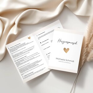 May include: A white card with the text "Herzensmensch" and a gold heart, with the name "Michaela Schmidt". The card is next to an open brochure with text. The background is a cream-coloured fabric.