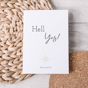 Könnte beinhalten: Weiße Karte mit dem Text "Hell Yes!" in schwarzer Kursivschrift. Unter dem Text befindet sich ein goldenes Sonnensymbol mit vier Strahlen. Unter dem Symbol steht der Text "#Bauchgefühl".