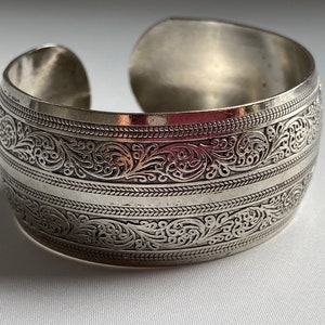 May include: A silver cuff bracelet with an intricate floral design. The bracelet is wide and has a rounded shape.