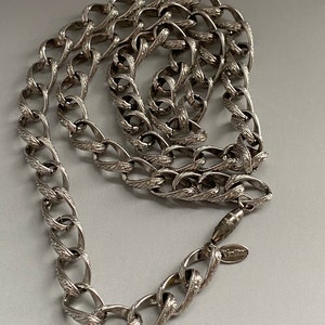 May include: A silver chain necklace with a detailed, ornate design. The chain is made of interlocking links with a textured surface. The necklace has a clasp with a small tag that reads "Vanities".