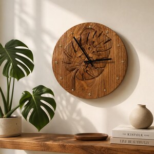 May include: A circular wooden wall clock featuring a carved monstera leaf design on its face. The clock has a natural brown wood grain finish, black hour and minute hands, and small gold-toned metal studs marking the hours around the perimeter.