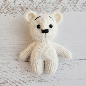 May include: A white knitted teddy bear with black eyes and a black nose. The bear is sitting on a white wooden surface.