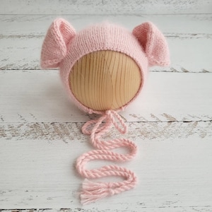Può includere: Un berretto per neonato di lana rosa con orecchie floppy e un lungo laccio. Il berretto è su una forma di legno.