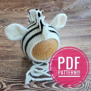May include: A knitted zebra hat with black stripes and white accents, featuring floppy ears and a fringe. The hat is placed on a wooden sphere. The image includes a pink circle with the text "PDF PATTERN!!!".