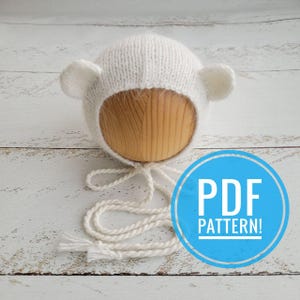 Pode incluir: Chapéu branco de tricô para bebê com orelhas de urso e uma gravata. O chapéu está em uma forma de madeira. O texto "PDF PATTERN!" está em um círculo azul.