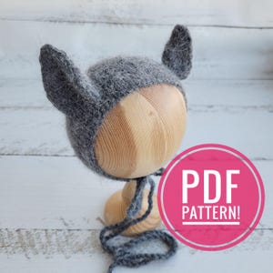 May include: A gray knitted baby hat with floppy ears, resembling a wolf or fox. The hat is displayed on a wooden mannequin head with a gray yarn tie. The image includes the text "PDF PATTERN!" in a pink circle.