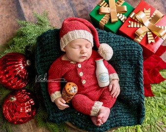 May include: A newborn baby wearing a red knitted Santa suit with a white pom-pom hat, holding a small milk bottle that says "Fresh Milk". The baby is lying in a green knitted basket with red and green Christmas ornaments and wrapped gifts nearby.