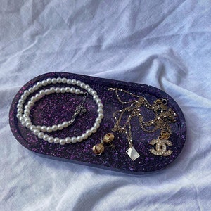 May include: A purple glitter oval tray with a pearl necklace, a gold chain necklace with a small white rectangular pendant, a gold chain necklace with a gold and crystal "C" charm, and a pair of gold earrings.
