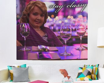 Puede incluir: Una mujer con cabello rubio y una camisa morada se sienta en una mesa con tres copas de vino. El texto "stay classy" está escrito en cursiva sobre su cabeza.