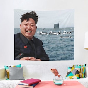 May include: A tapestry featuring a portrait of Kim Jong Un with the quote "Live, Laugh, Love" - Kim Jong Un. The background is a blue ocean with a submarine in the distance.