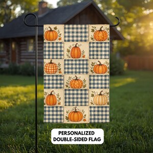Pode incluir: Uma bandeira de jardim retangular com um design de patchwork de abóboras cor de laranja e quadrados axadrezados azuis e brancos. A bandeira é exibida num suporte de metal preto e inclui o texto PERSONALIZED DOUBLE-SIDED FLAG na base.