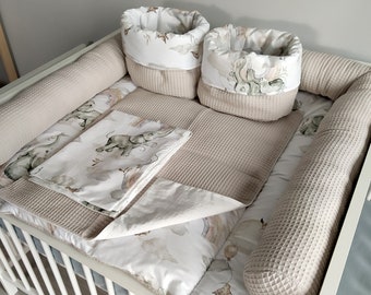 May include: A white crib with a beige and white baby bedding set. The bedding includes a fitted sheet, a blanket, two bumper pads, and two storage baskets with elephant designs.