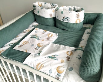 May include: A green and white baby crib bedding set with a hot air balloon and airplane theme. The set includes a bumper, a fitted sheet, a blanket, and two storage baskets.
