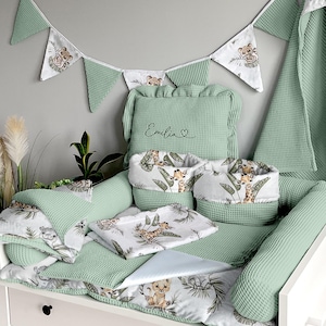 Puede incluir: Un conjunto de decoración de guardería verde y blanco con un parachoques de cuna, una almohada y una manta. El conjunto presenta un estampado de animales lindo con una jirafa y un león. La almohada tiene el nombre "Emilia" bordado.
