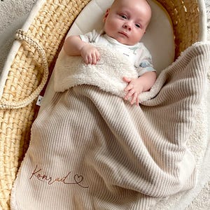 Könnte beinhalten: Ein Baby liegt in einem geflochtenen Korb, bedeckt mit einer beigefarbenen Decke. Die Decke hat ein weiches, flauschiges Futter und den Namen "Konrad" in Schreibschrift gestickt. Der Korb steht auf einem runden Teppich mit Fransen.
