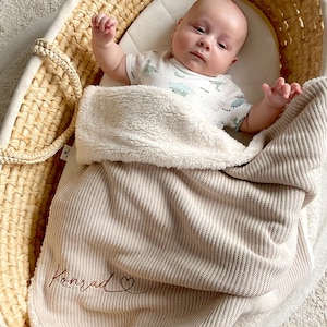 May include: A baby lies in a woven bassinet, covered by a beige blanket embroidered with the name "Konrad". The blanket has a soft, plush lining. The bassinet rests on a round rug with a fringed edge.