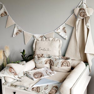 Può includere: Un set per la cameretta con un fasciatoio bianco e tessuti beige. Il set include un fasciatoio, cuscini, un asciugamano con cappuccio e una ghirlanda. I tessuti presentano disegni di gufi e animali del bosco. Il nome "Emilia" è ricamato su un cuscino.