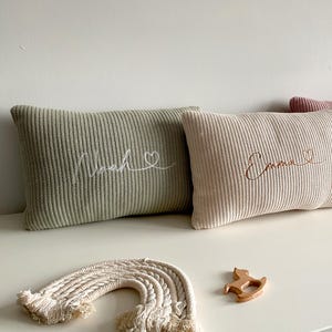 May include: Three rectangular knit cushions in sage green, cream, and dusty rose. Each cushion features embroidered cursive names: Noah, Emma, and a third name, accompanied by a small heart outline. The cushions have a ribbed texture.