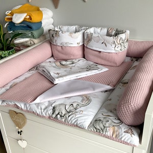 Peut inclure: Un matelas à langer blanc avec un imprimé d'éléphants rose et blanc. Le matelas est entouré de pare-chocs en gaufre rose et de deux paniers de rangement rose avec des imprimés d'éléphants blancs et gris. Le matelas à langer est sur une commode blanche avec un cœur en bois qui dit "Love" suspendu à celui-ci.