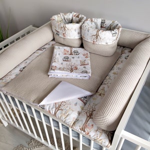 Peut inclure: Un lit bébé blanc avec un pare-chocs de lit beige et blanc, un matelas à langer en tissu gaufré beige, un matelas à langer blanc et deux paniers de rangement beige et blanc avec un imprimé d'animaux de la forêt.