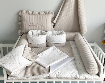 Könnte beinhalten: Ein beige-weißes Babybett-Bettwäsche-Set mit Waffelstruktur. Das Set beinhaltet einen Stoßfänger, ein Spannbettlaken, eine Decke, ein Kissen und zwei kleine Körbe. Das Kissen ist mit dem Namen "Paul" in Rot bestickt.