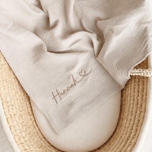 Peut inclure: Une couverture d'emmaillotage en mousseline beige clair avec une texture froissée, présentant le prénom Hannah brodé suivi d'un petit cœur en fil marron foncé, posée dans un couffin ovale en osier tressé avec des anses tressées.