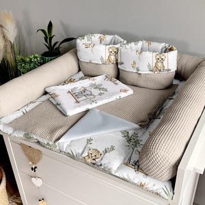 Op de afbeelding: Een beige verschoningsmat met een wit en groen gepatroneerd, gestikt inzetstuk. Twee beige wafelstructuur verschoningsmat bumpers bevinden zich aan beide zijden van de mat. Twee wit en groen gepatroneerde opbergmanden met dierillustraties bevinden zich op de mat.