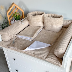 May include: A beige baby changing station set on a white dresser. The set includes a changing pad, two bolster pillows, two storage baskets, and a small towel. The items are made of waffle-textured fabric.
