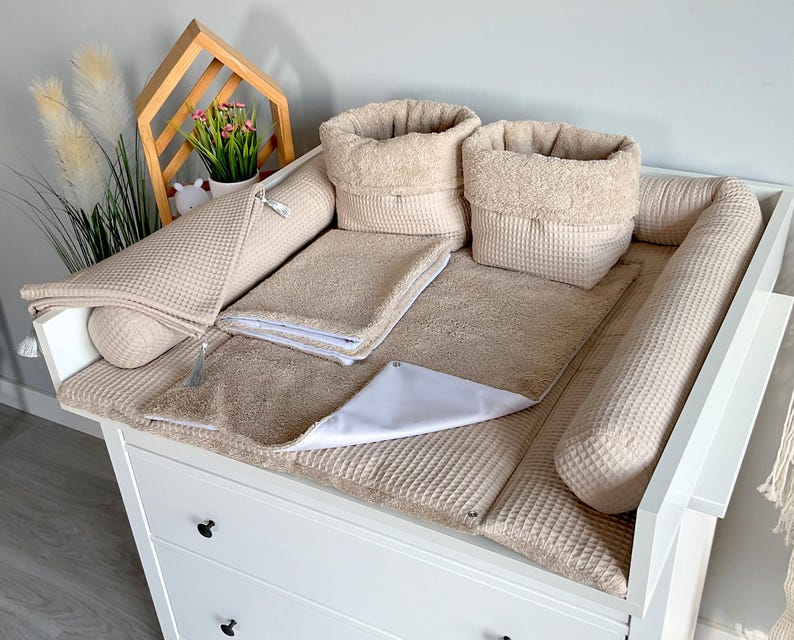 May include: A beige baby changing station set on a white dresser. The set includes a changing pad, two bolster pillows, two storage baskets, and a small towel. The items are made of waffle-textured fabric.