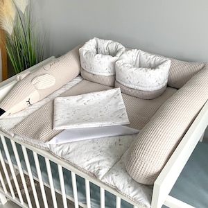 Könnte beinhalten: Ein weißes Babybett mit einem beigefarbenen, gewellten Stoßfänger und einem beige-weiß gemusterten Spannbettlaken. Das Bett enthält auch ein beige-weiß gemustertes Kissen und ein beigefarbenes, gewelltes Stofftier in Form eines Bären.